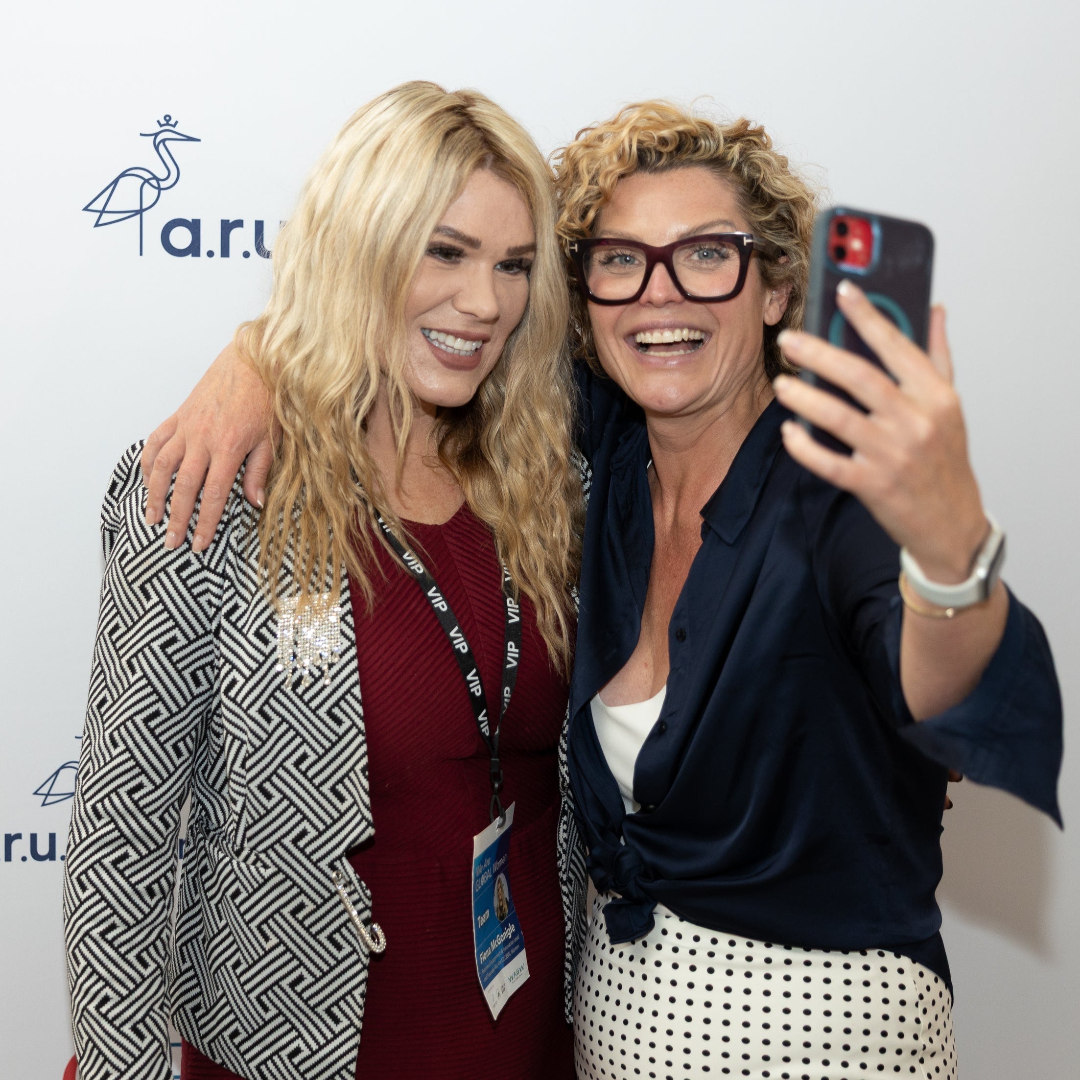Two women taking a selfie with a phone, wearing lanyards, in front of a white background with 'a.r.' logo.