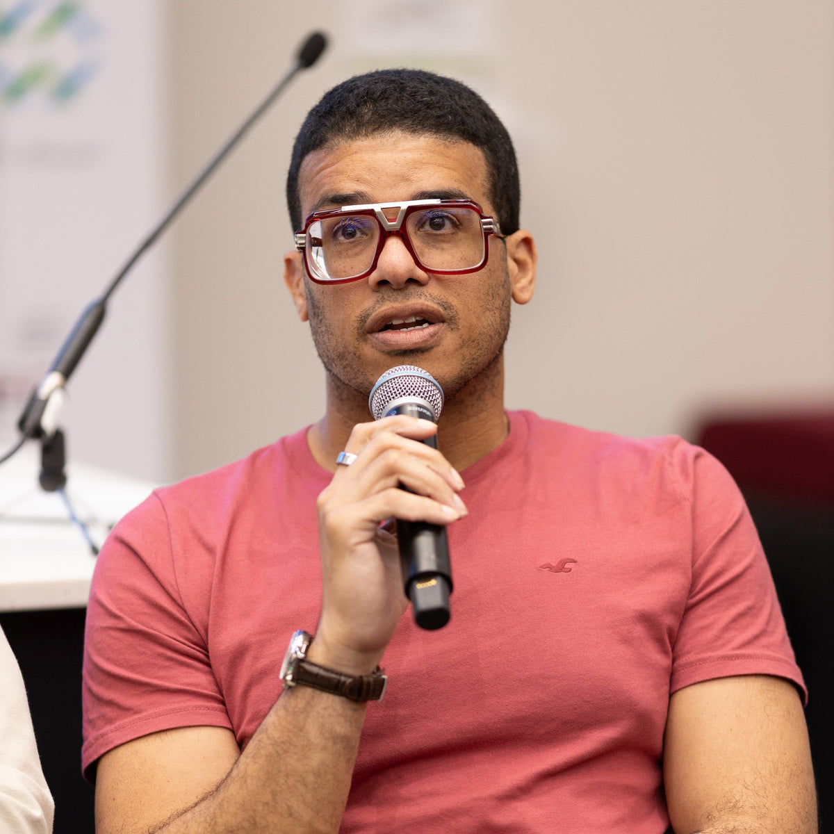 Man in a pink shirt holding a microphone, wearing red glasses.