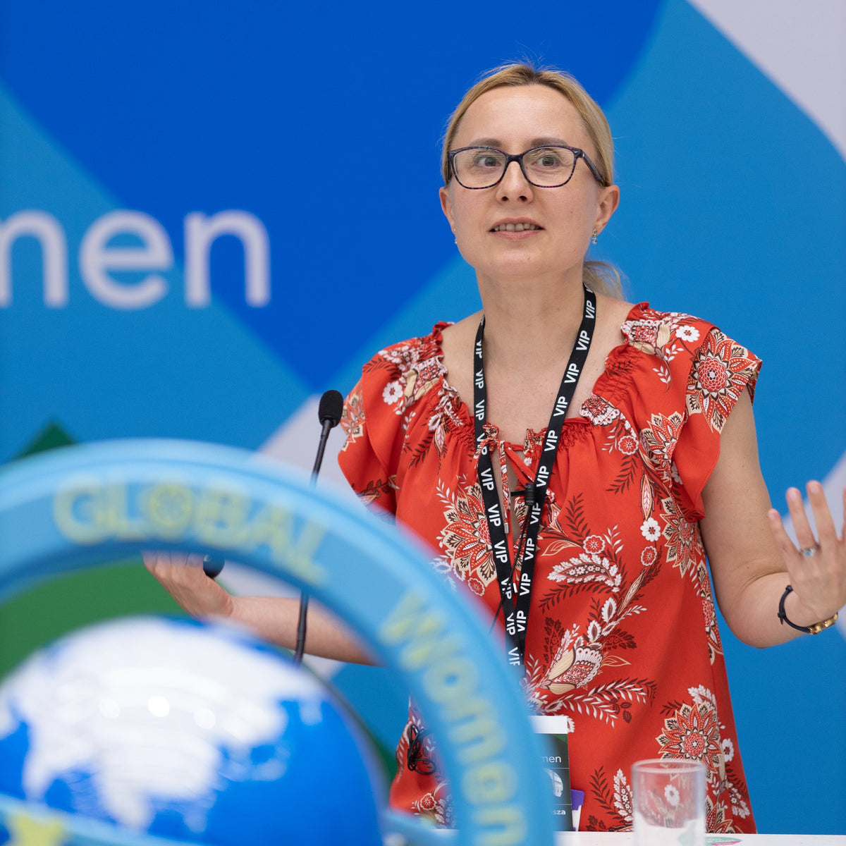 Woman in a red floral blouse speaking at an event with a blue background