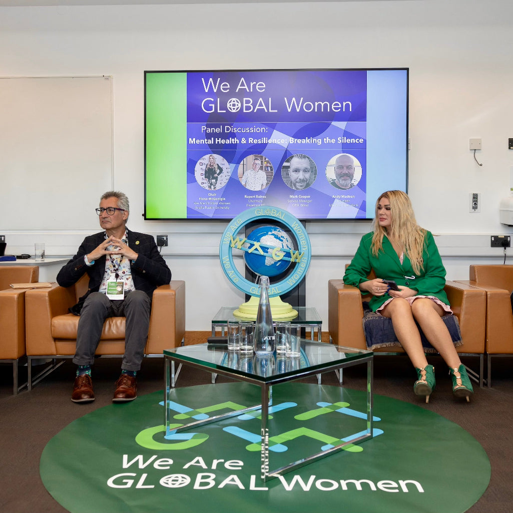 Group of people sitting on a stage with a screen displaying 'We Are GLOBAL Women' in a conference setting.